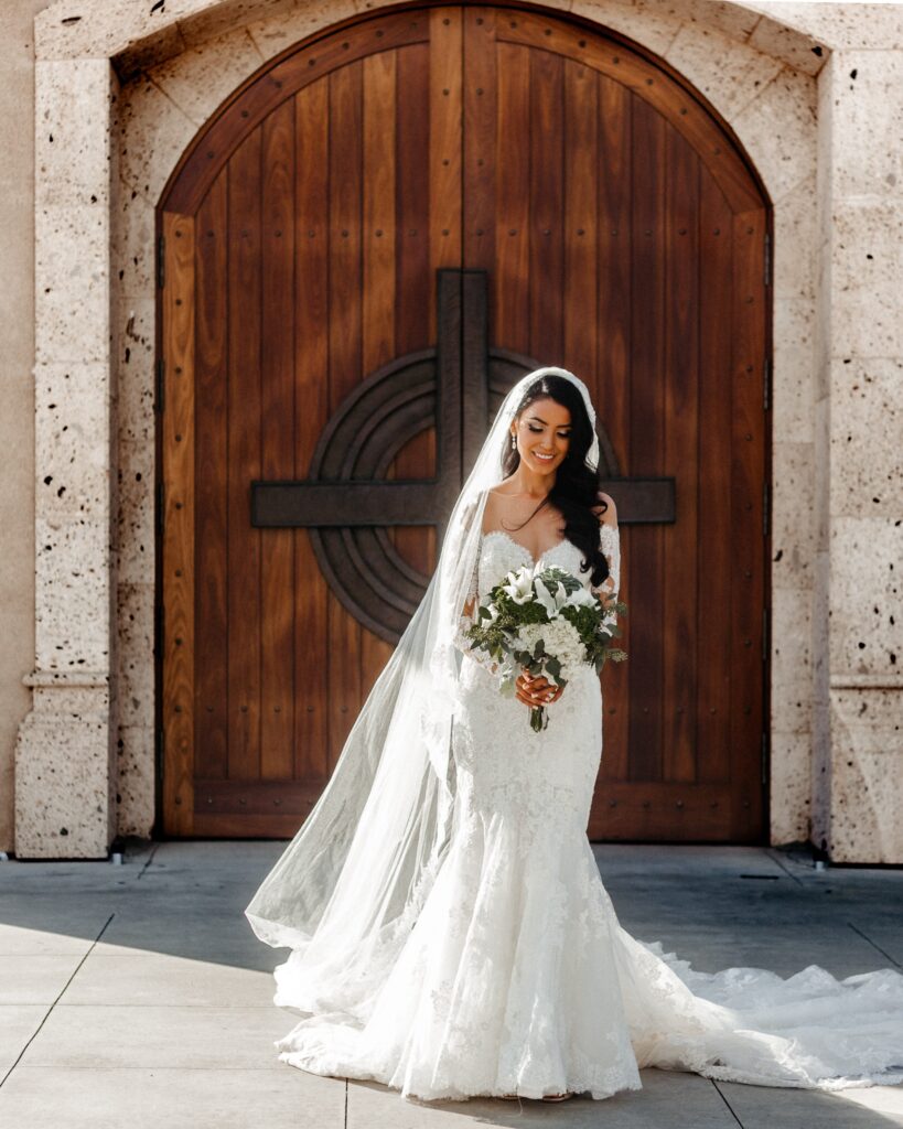 tan bride standing in front of a church holding a bouquet